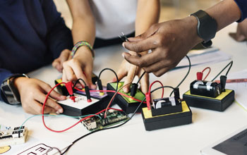 one hand holding tweezers while 2 others building electronics. 36 black and red wires plugged in to boards