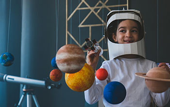 boy wearing cardboard astronaut helmet flying toy rocket through planets, cardboard spaceship rocket in the background
